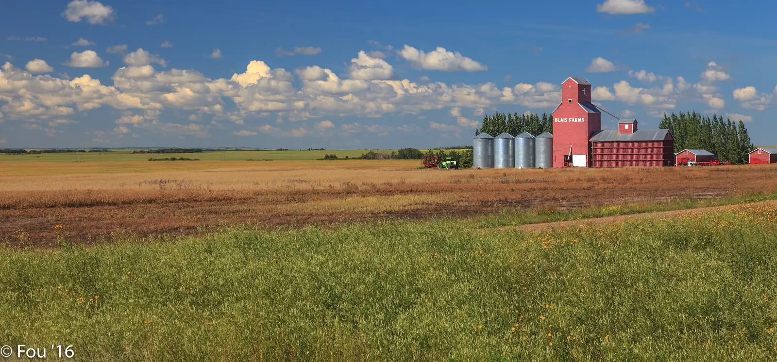 A red wooden grain elevator and steel bins beside a harvested field under a big blue Saskatchewan sky